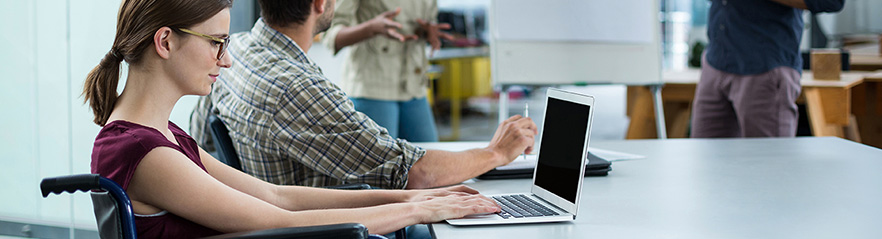 A women in a wheel chair seated at a table in a conference room using a laptop computer while other team members talk.