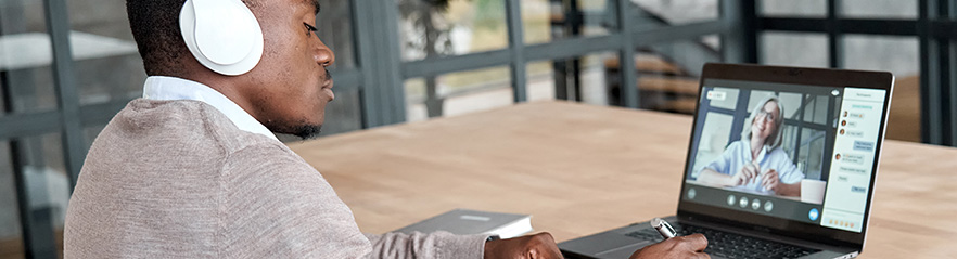 A man wearing headphones sits at a table and uses a laptop computer for a video meeting