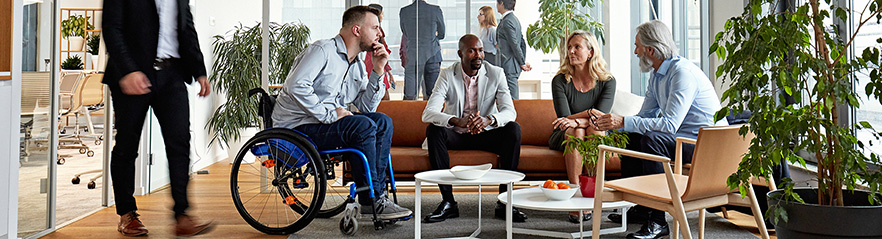 A group of diverse people sitting on a couch, chair, and in a wheelchair are engaged in conversation around a table.