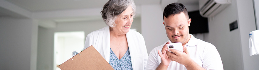 A clinician in a white coat holding a clipboard observes a smiling young man as he looks down at his cell phone.