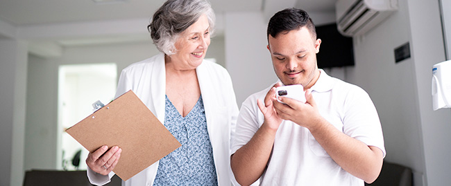 A clinician in a white coat holding a clipboard observes a smiling young man as he looks down at his cell phone.