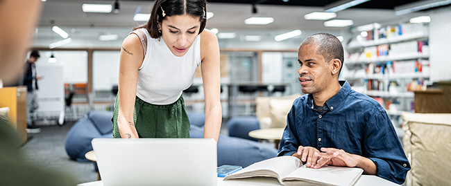 A man in a library room is reading from a braille book, and a women at the same table looks at a laptop computer screen.