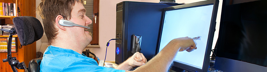 A man in a wheelchair with a microphone headset on uses a computer touch screen