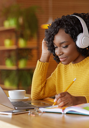 A women seated at a desk looks at a laptop screen while wearing headphone and holding a pencil to take notes