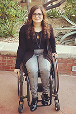 Toni Saia, a light-skinned female wheelchair user with round tortoise-shell glasses and long brown hair, wearing a black scoop-necked shirt, black jacket and grey pants.